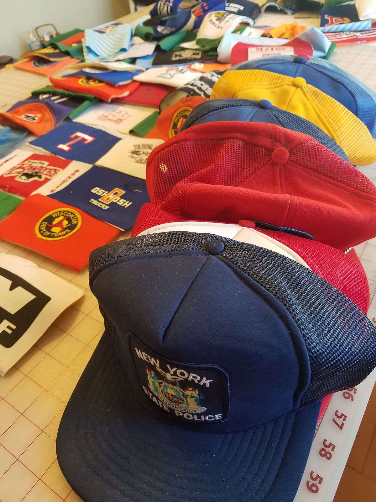 Quilt squares feating different sports team logos lay out on a cutting board next to a row of baseball caps.