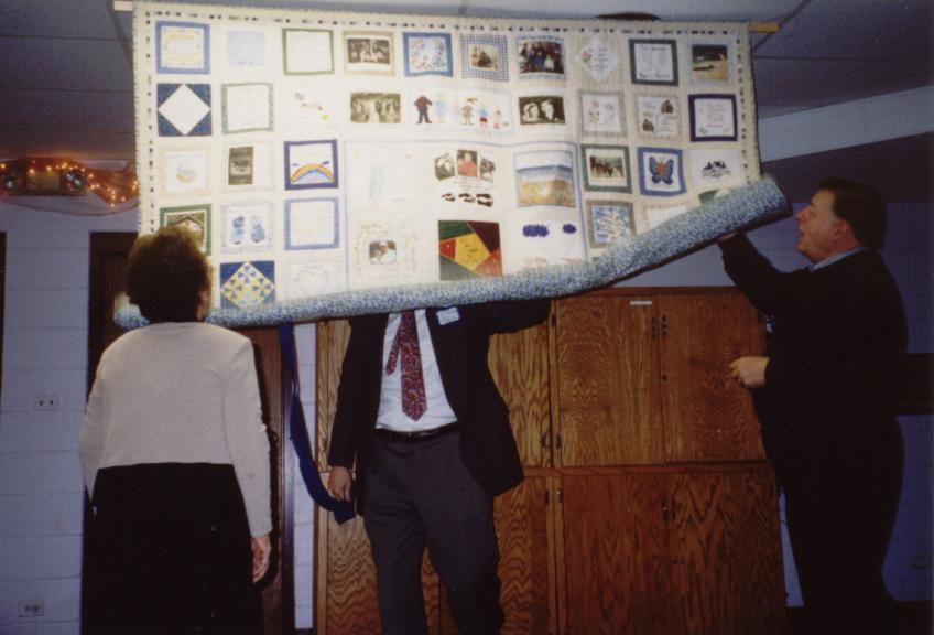 A retired couple watch as a suprise anniversary quilt is unrolled for them, made with squares from each member of their family.